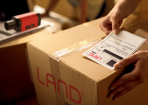 A LAND-branded shipping box being labelled for dispatch, with a LAND thermal imaging camera visible in the background.