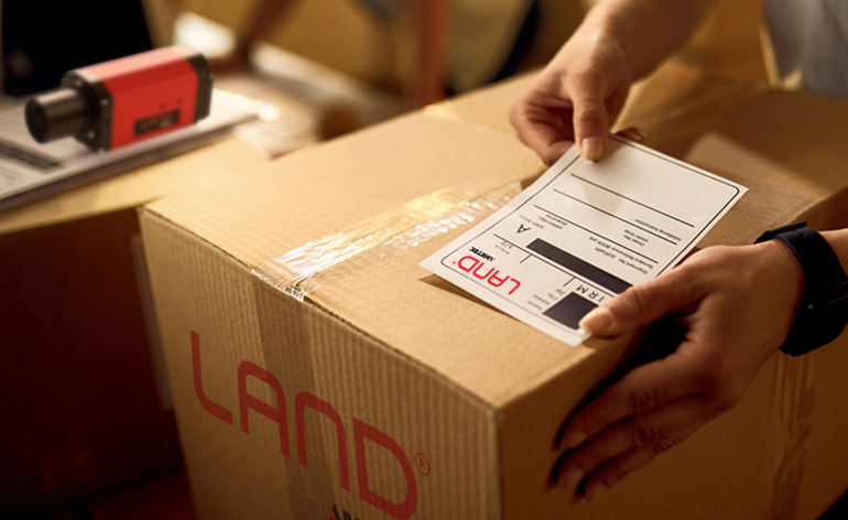 A LAND-branded shipping box being labelled for dispatch, with a LAND thermal imaging camera visible in the background.